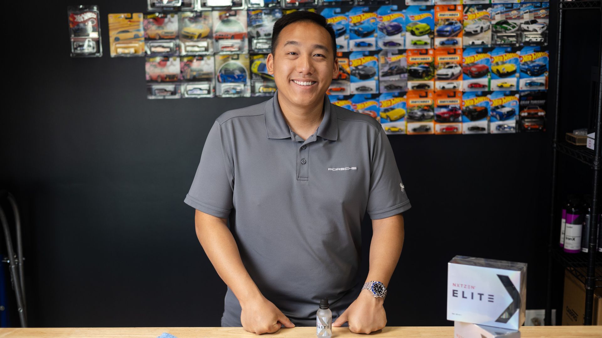 A smiling individual wearing a gray polo shirt stands behind a table with a box in a room featuring walls lined with toys.