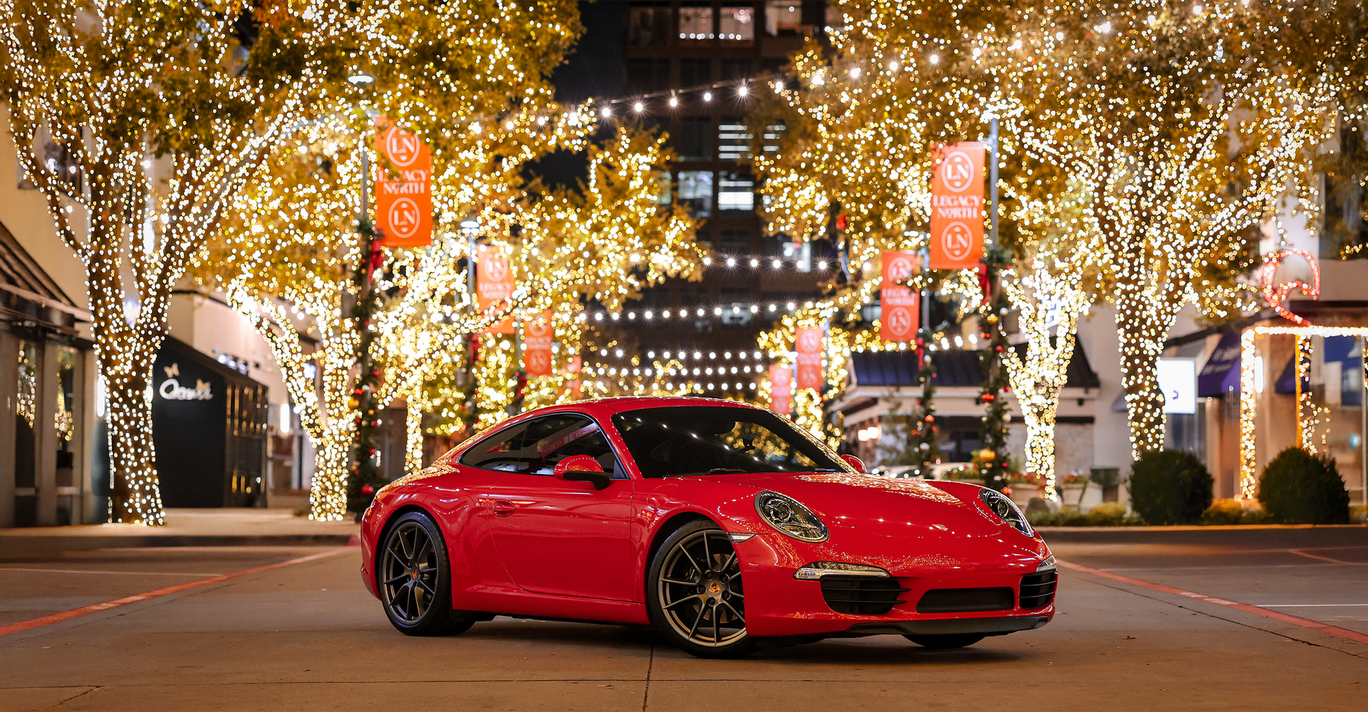 A bright red Porsche parked on a city street at night, surrounded by trees illuminated with warm, festive string lights.