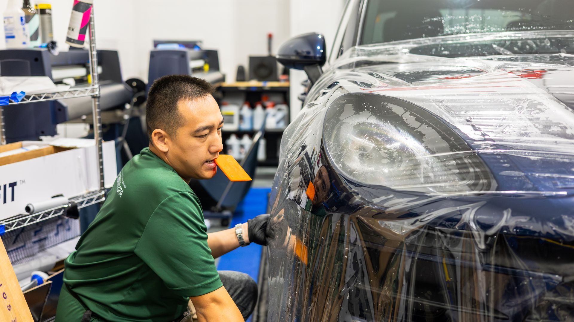Mechanic in green shirt wraps black car with protective film in an auto shop