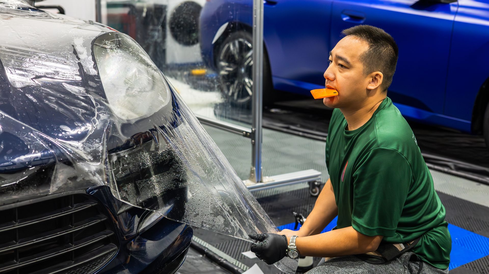 Man in green shirt crouches beside a car front covered in plastic wrap in an auto shop.