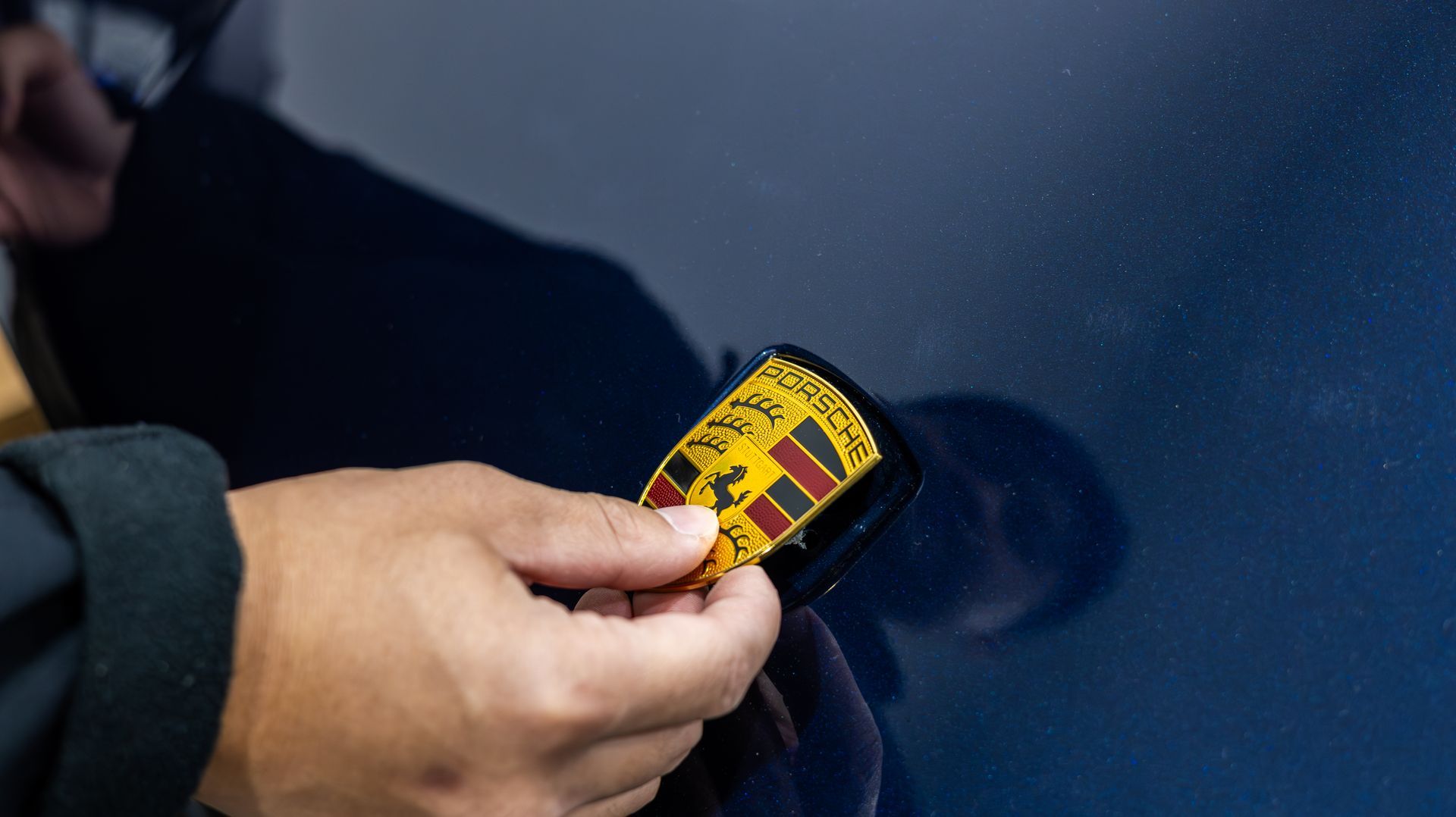 Hand holding a yellow badge against a dark blue car door, showing a small dent or scratch