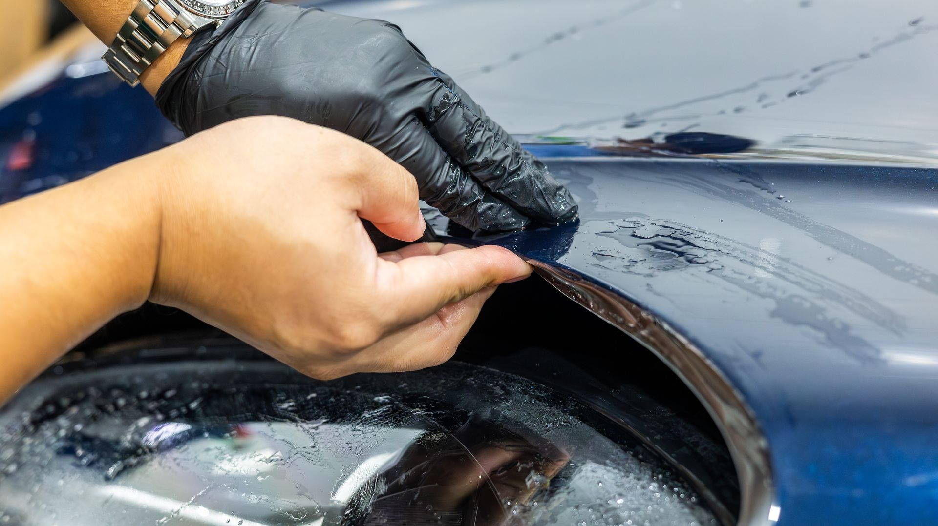 Hands polishing a blue car’s headlight with a cloth and glove