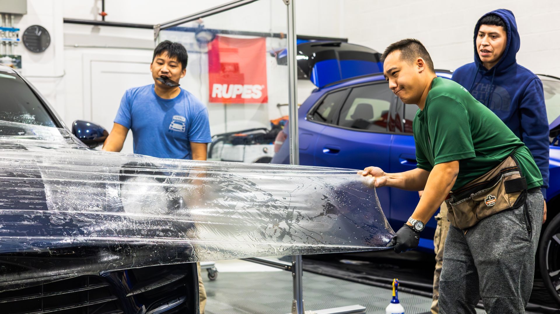 Workers washing a black car in a garage, spraying foam and wiping the hood.