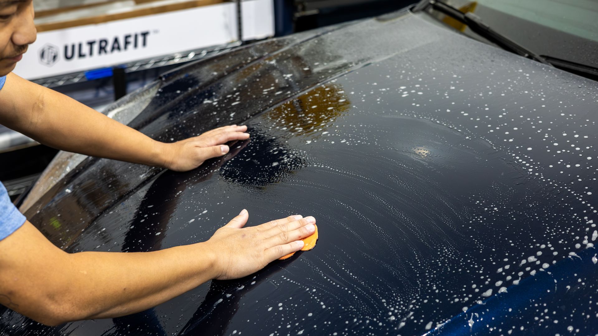 Hands using a sponge to wash a black car covered in soap suds.