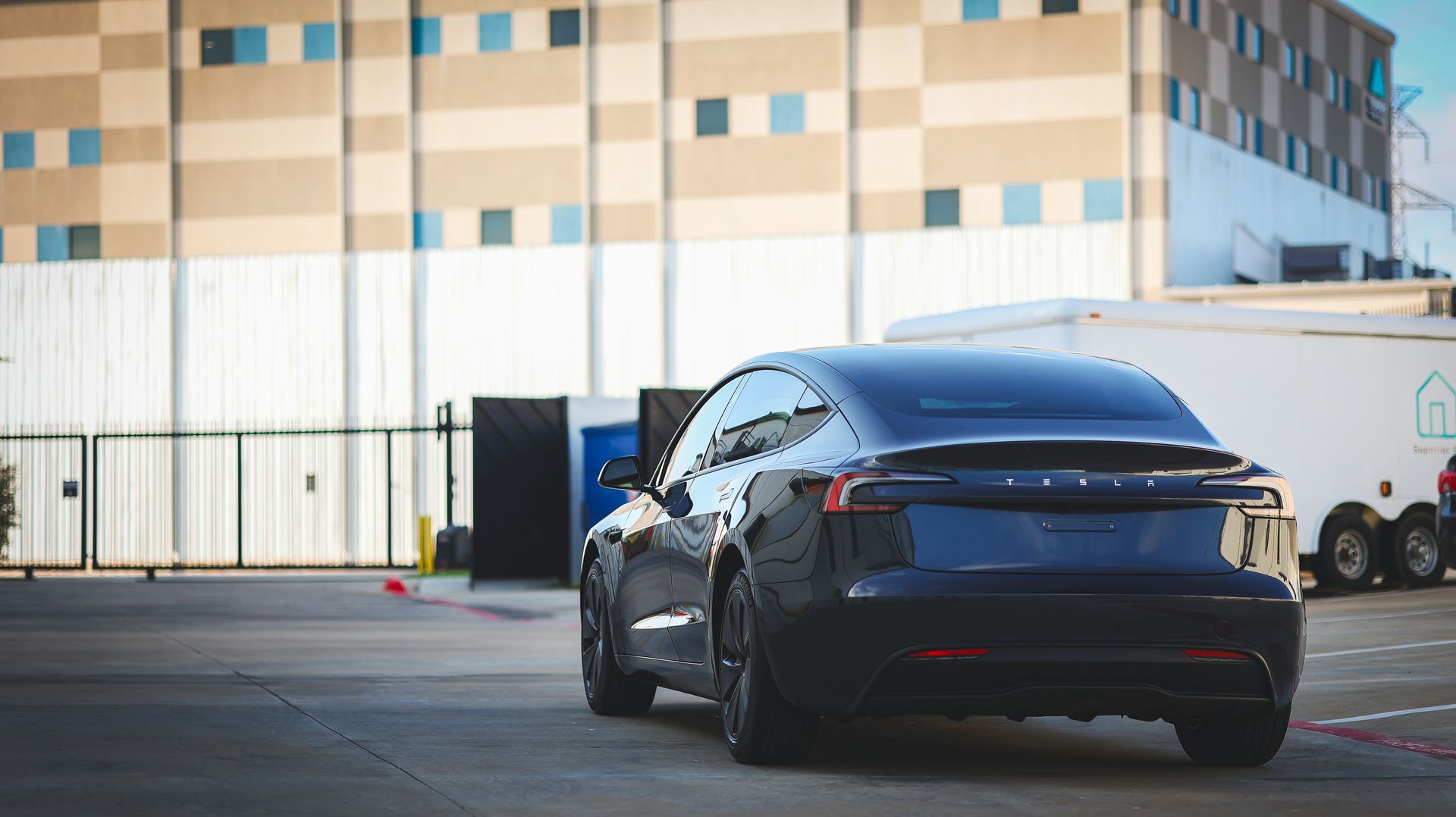 A dark blue Tesla Model 3 parked in a paved lot in front of a tan industrial building with a white trailer to the side.