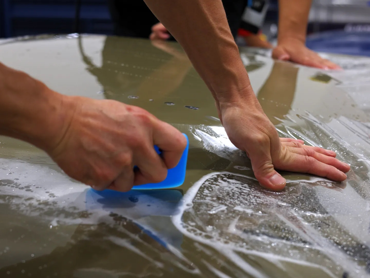 Installer applying paint protection film to vehicle hood using squeegee to remove air bubbles and water