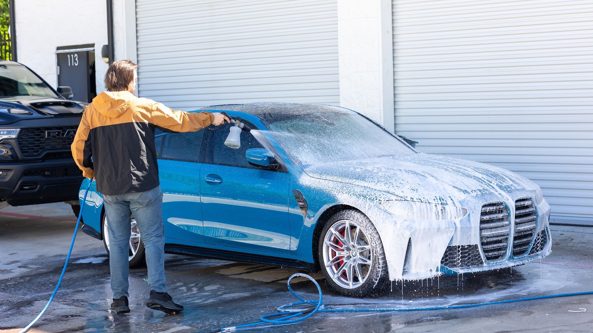 Person washing a blue sedan with a hose in a car wash bay, covered in soap suds.