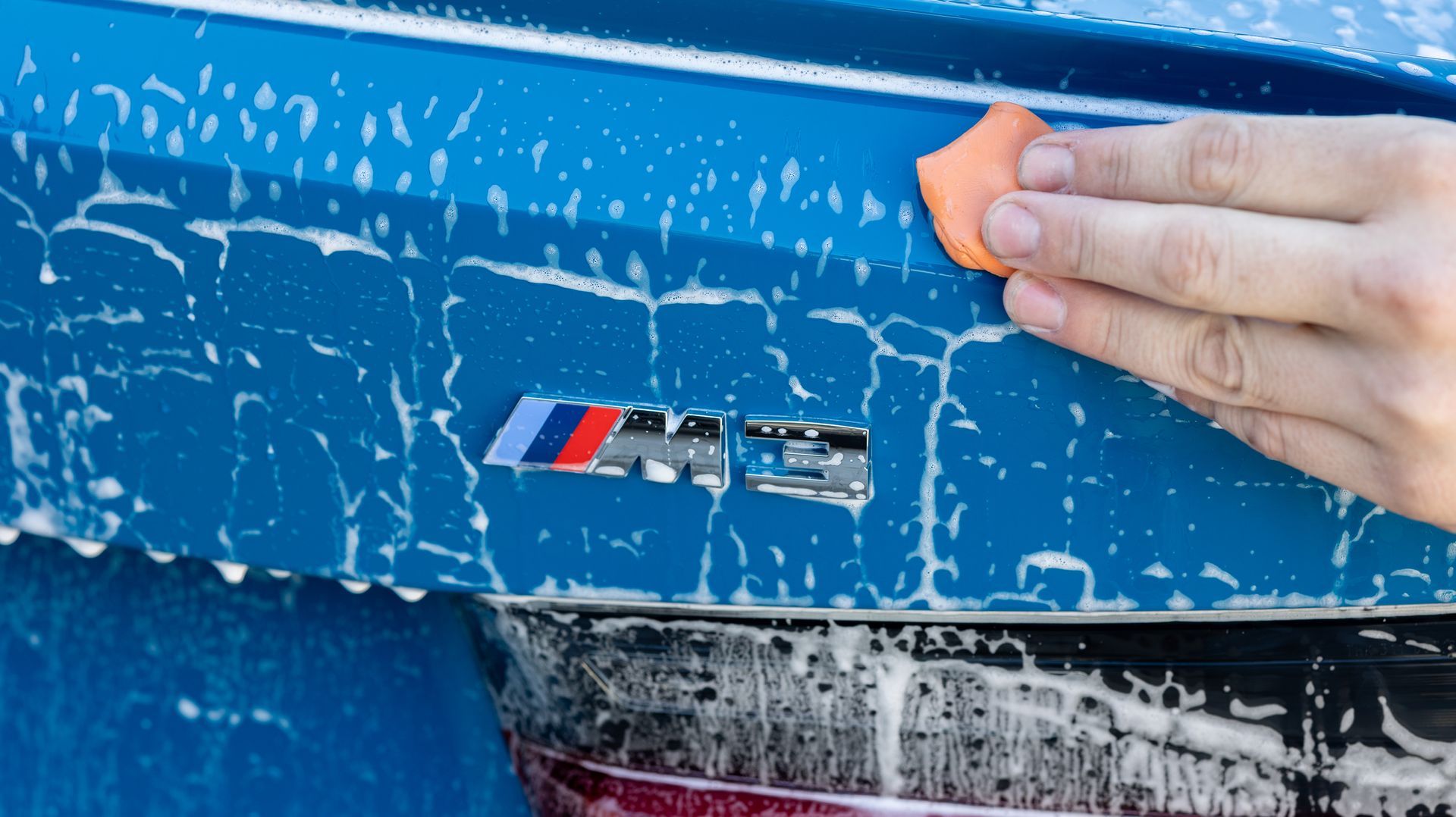 Hand rubbing mud off a blue car panel, revealing an M badge.