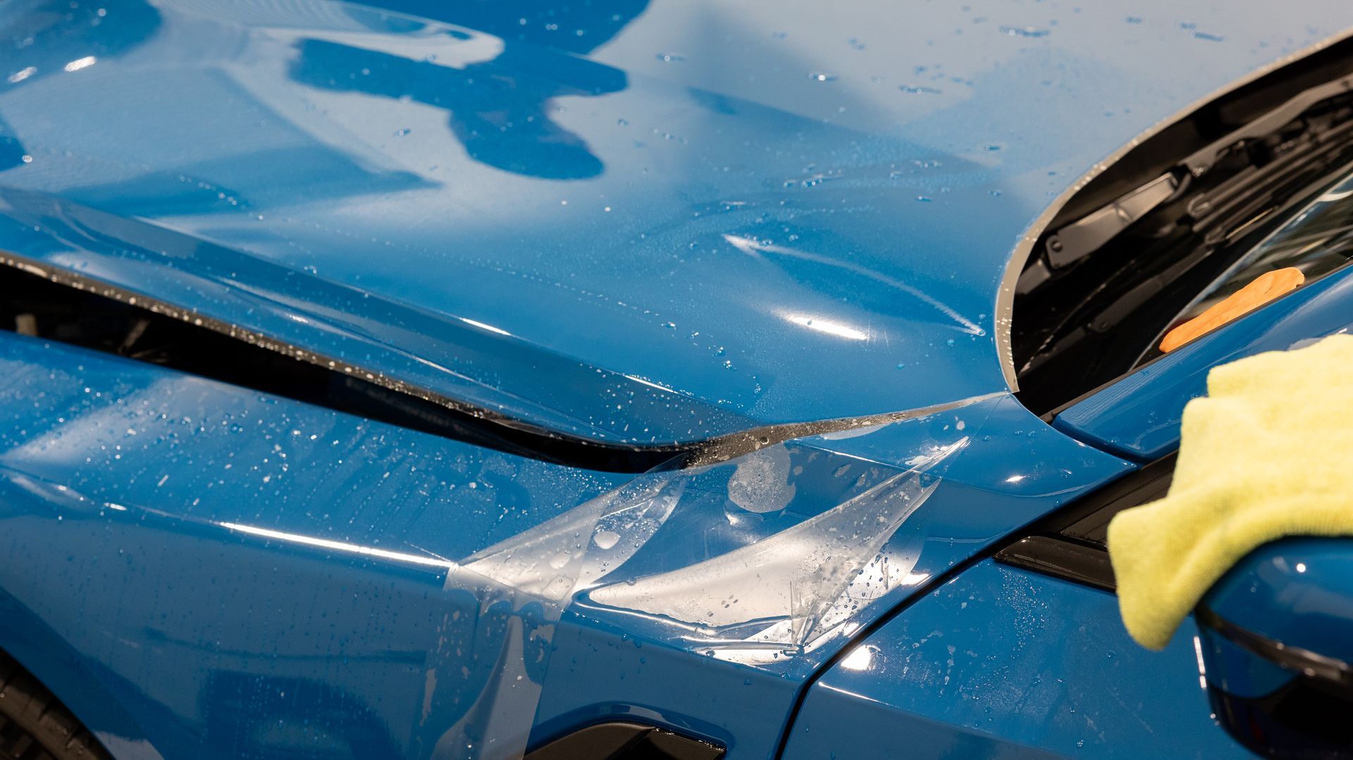 Blue car being washed by a person wearing yellow gloves, with soap and water on the hood