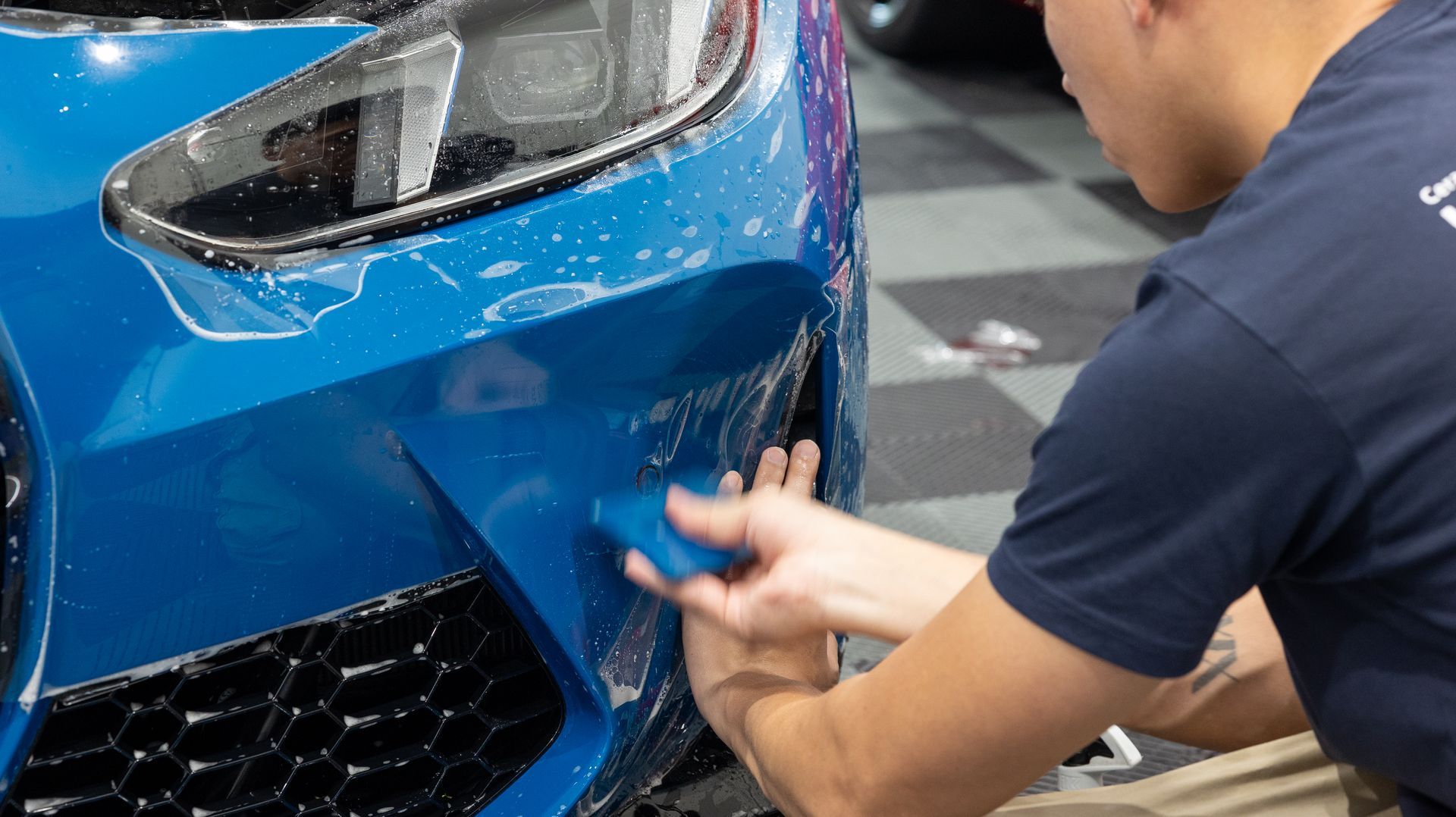 Person polishing the front bumper of a blue sports car in a workshop