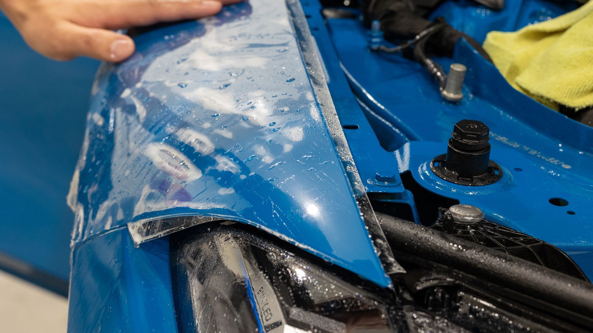 Hand peeling a blue protective film from a car body panel near exposed engine parts