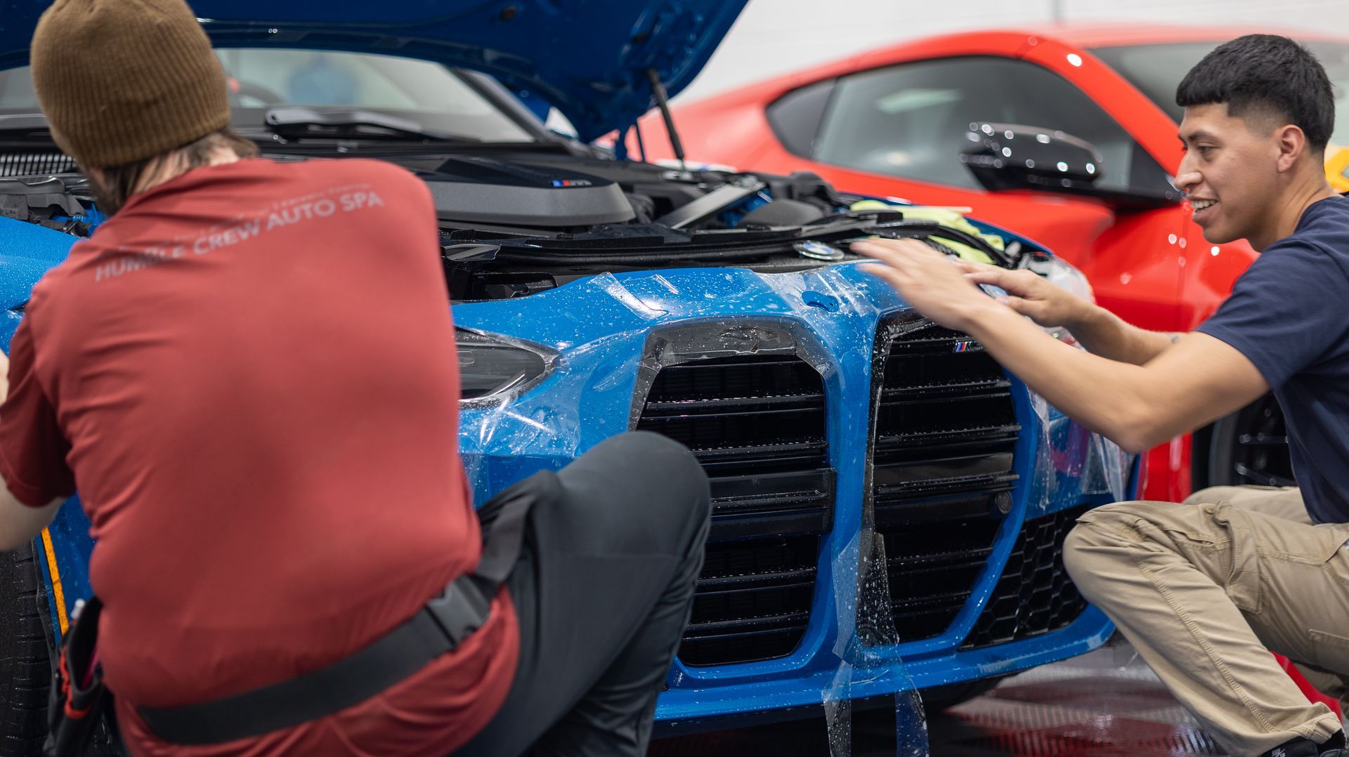 Two people work on the front of a blue sports car at an auto body shop.
