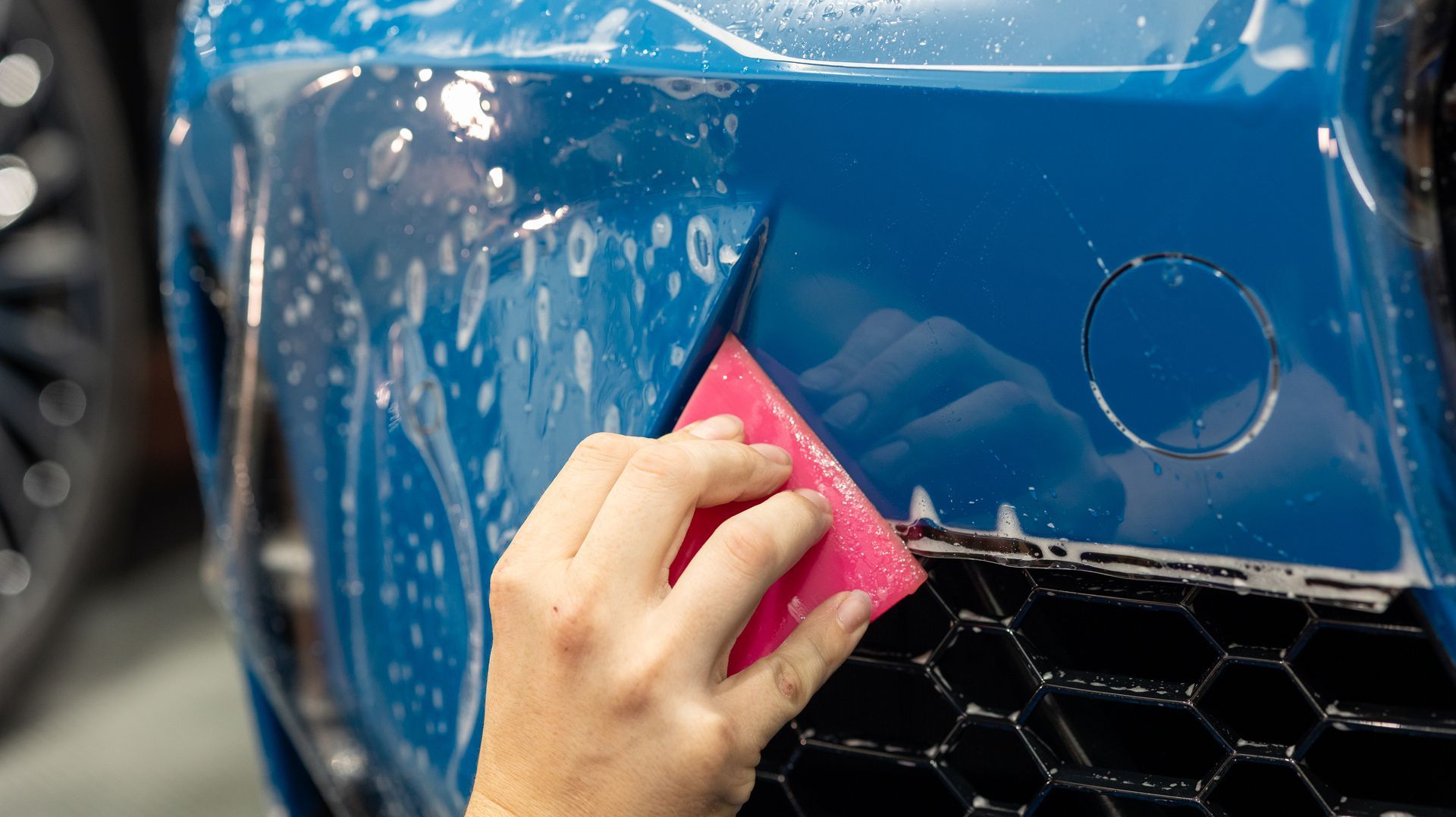 Hand cleaning a blue car’s front bumper with a pink cloth