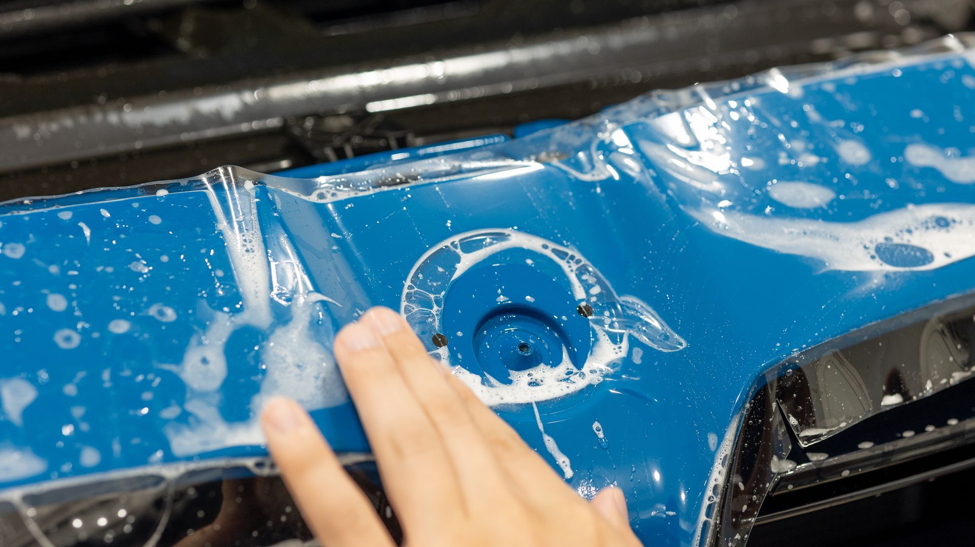 Hand washing a blue car’s door, with soap suds on the paint and a fuel cap visible