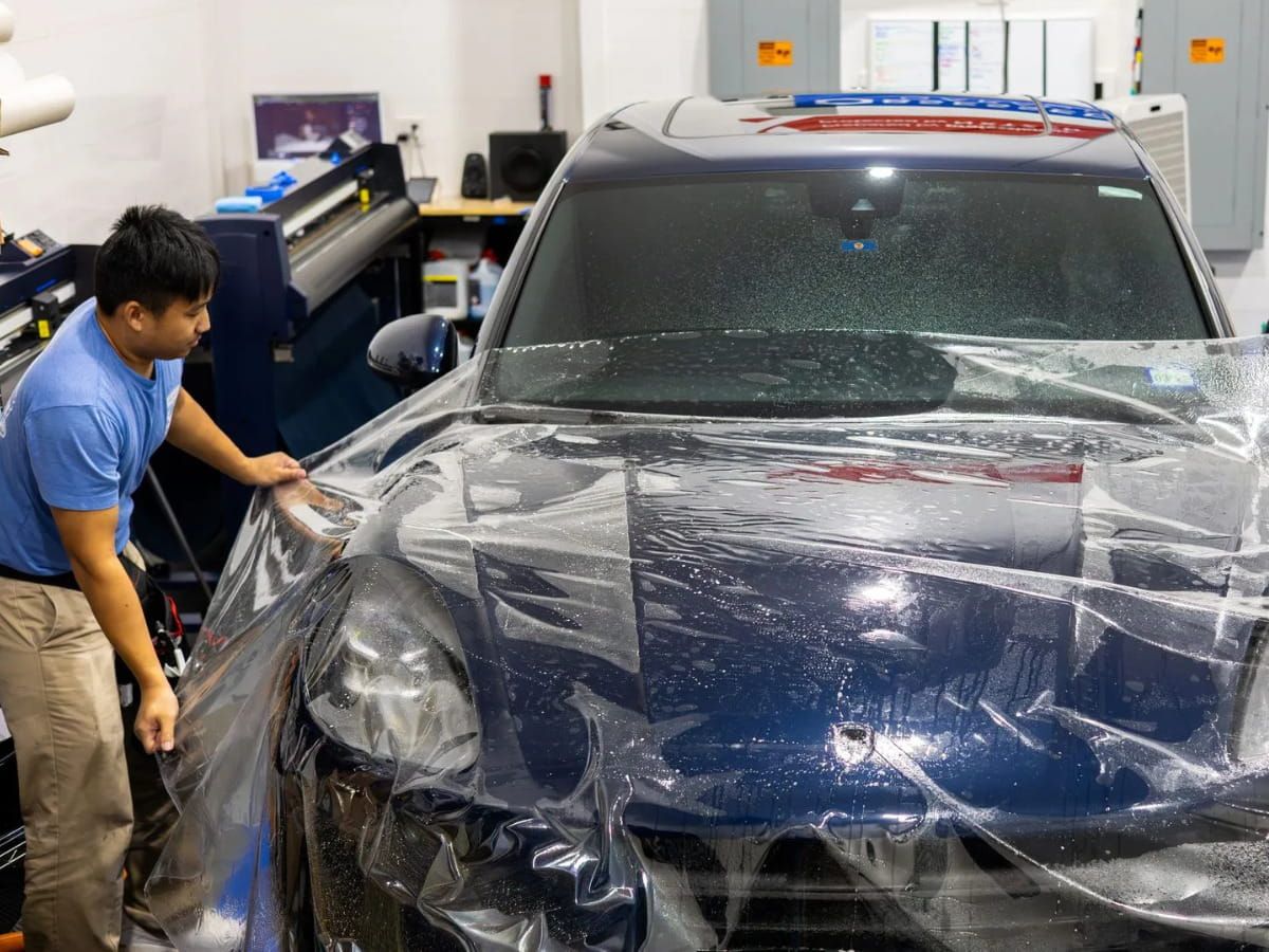 Technician installing paint protection film across the hood and front fender of a blue vehicle.