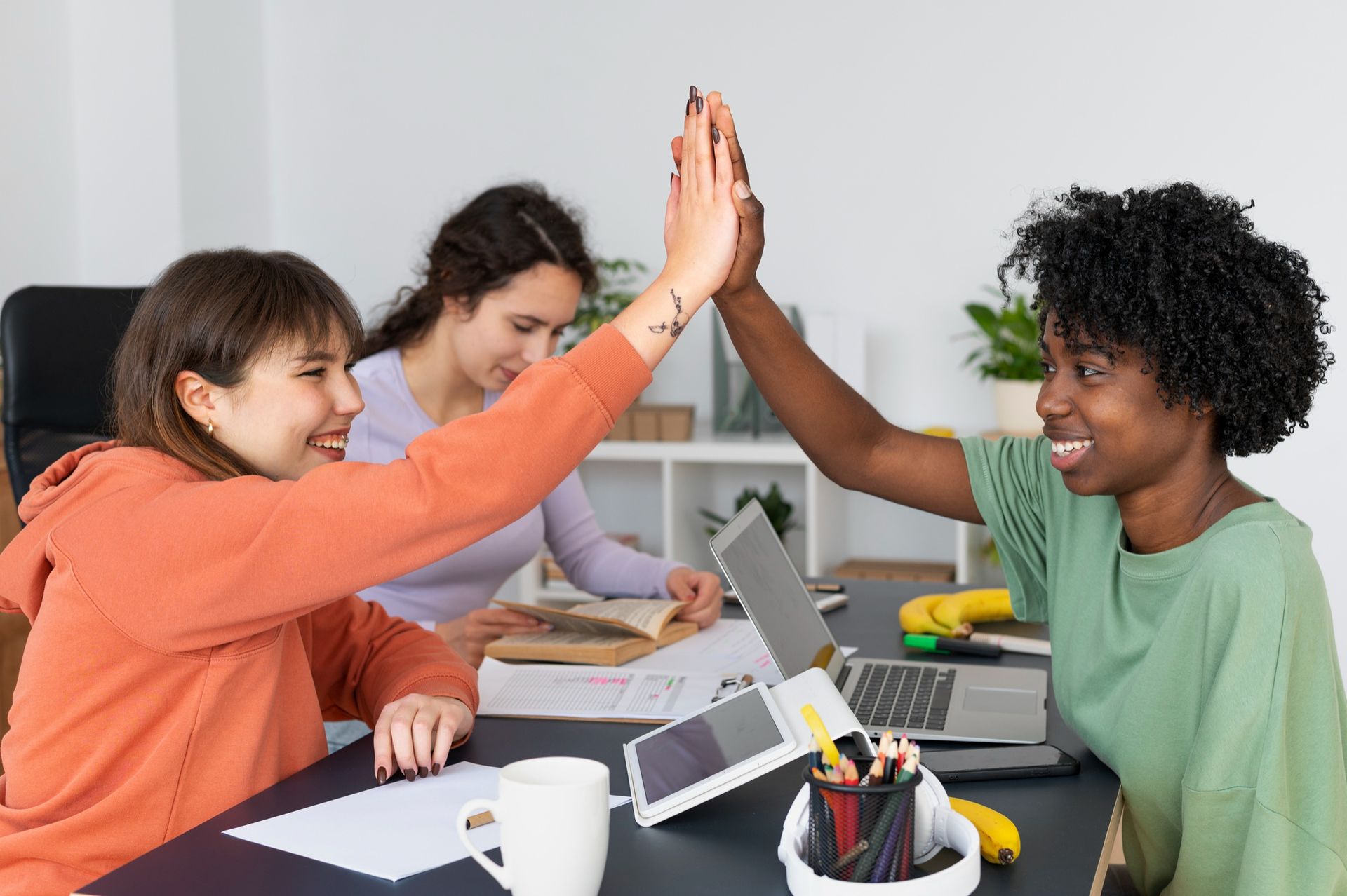 People high-five across a table in a bright meeting room, with laptops, papers, and a coffee cup nearby.