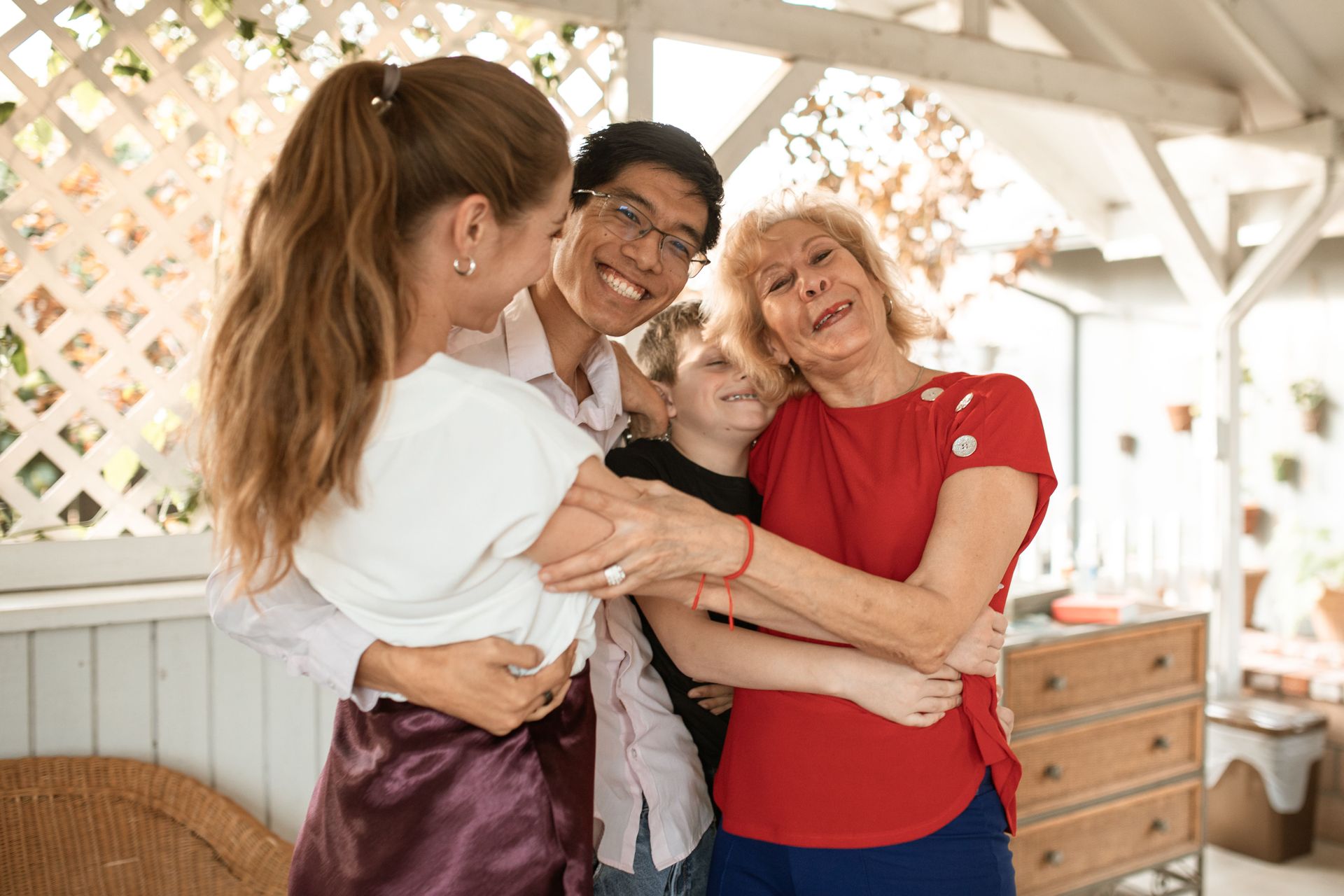 Four people hugging and smiling in a bright sunroom with white lattice and wooden furniture