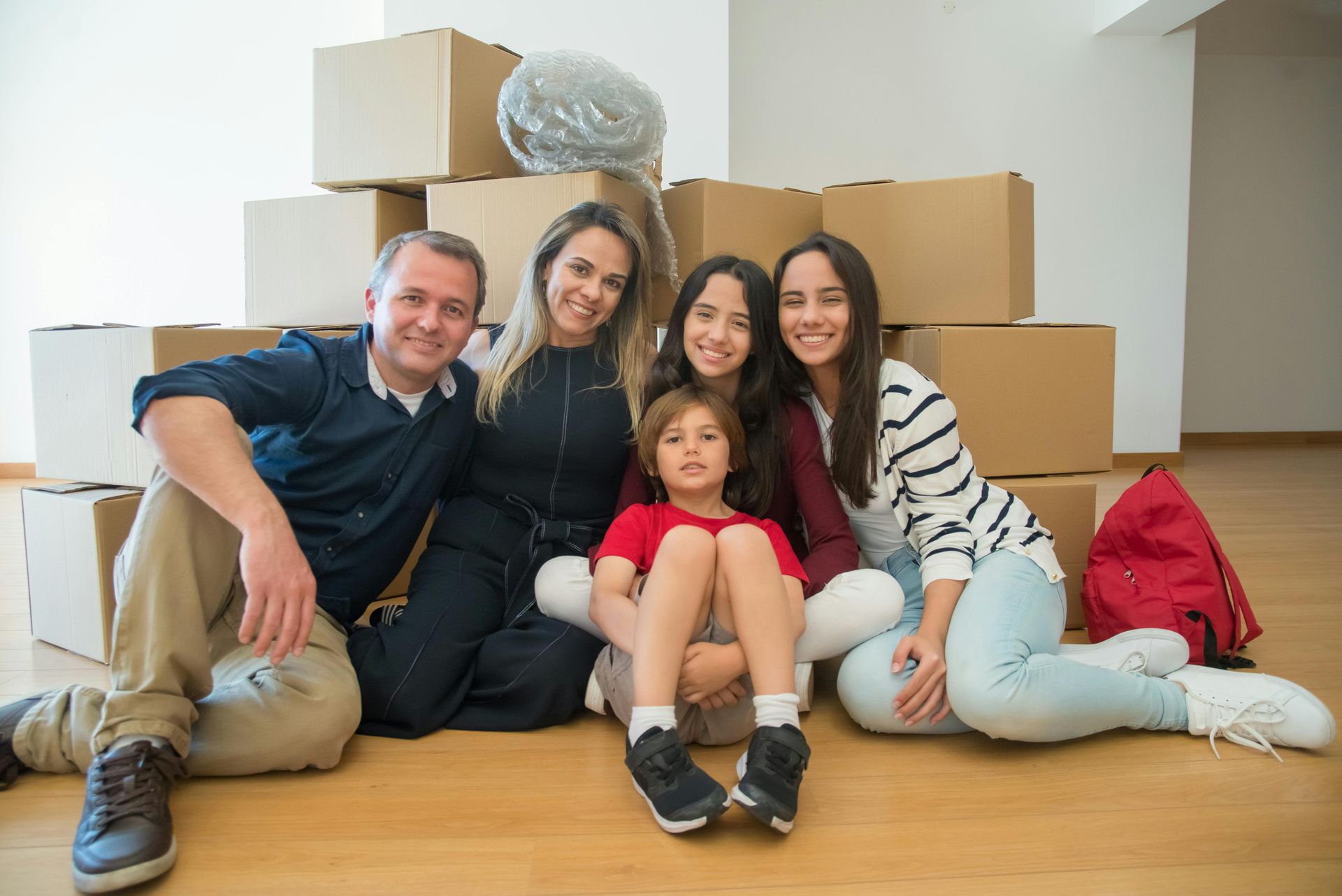 Family sitting on a wooden floor with moving boxes in a bright room, smiling at the camera