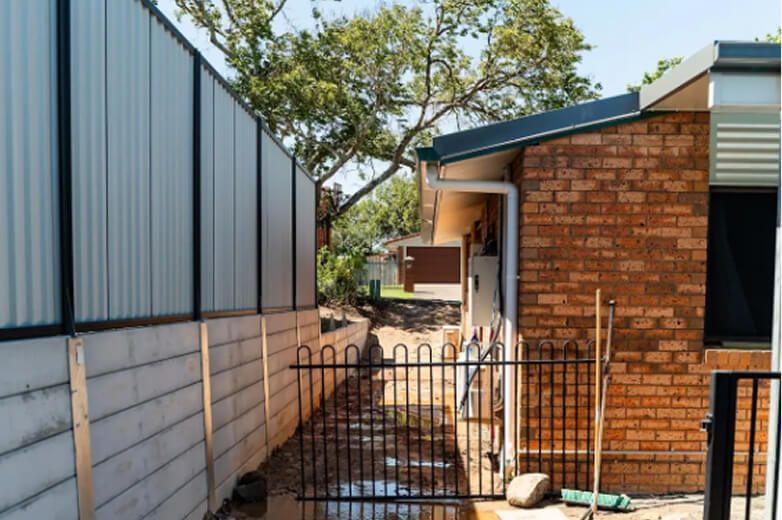 Narrow side yard with metal fence, brick house, and tree in daylight