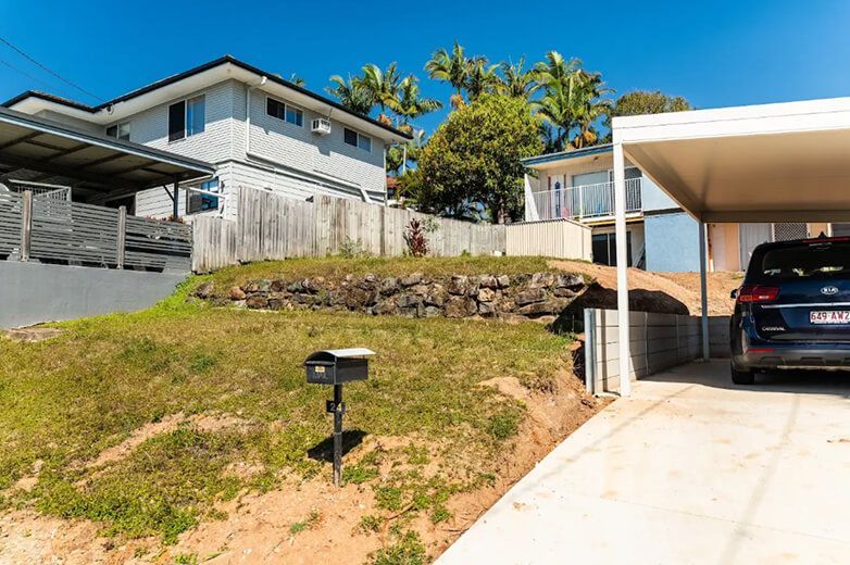 Residential driveway beside a modern house, with a car under a white carport and a grassy slope with a mailbox.