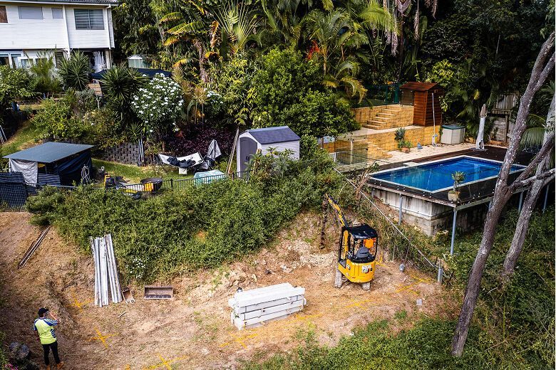 Backyard construction site with a pool, small sheds, a yellow excavator, and stacked materials.
