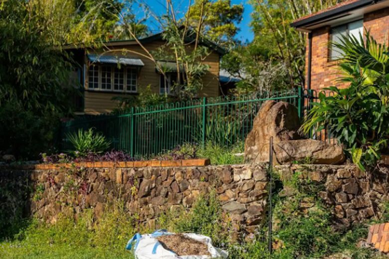 Stone retaining wall and green fence in front of a house with trees and bushes in a sunny garden area