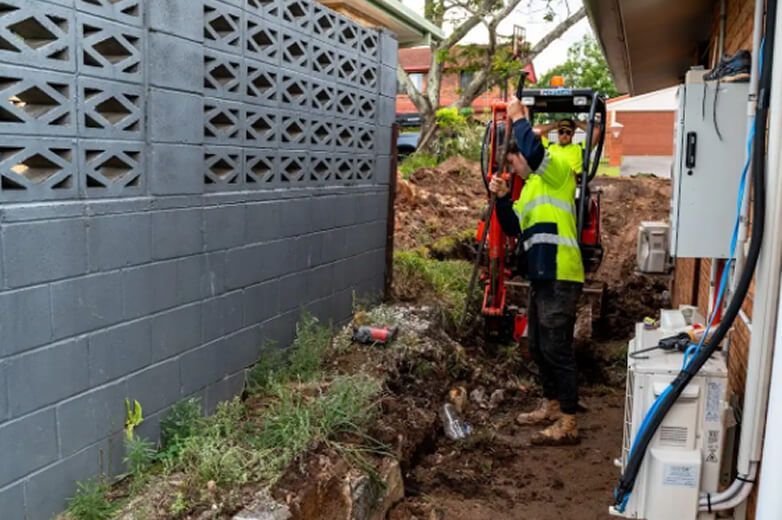 Workers in neon vests digging a narrow trench beside a house and gray block wall