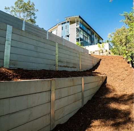 Curved concrete retaining walls beside a dirt path on a hillside, with a modern building above.