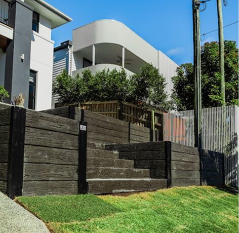 Modern house with white curved balcony above dark timber retaining walls and green lawn under a clear blue sky