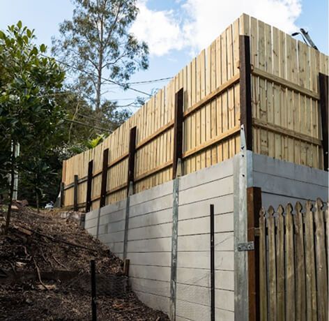 Tan retaining wall beneath a tall wooden fence on a wooded hillside.