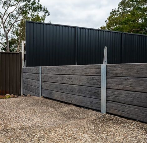 Modern black metal fence above a gray timber retaining wall beside a gravel driveway