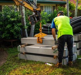 Worker in neon shirt using a crane to lift concrete slabs in a yard.