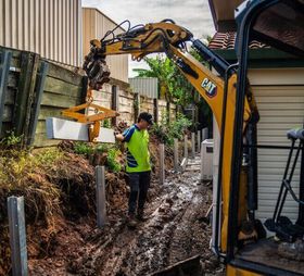 Worker guiding a CAT excavator beside a muddy trench and fence in a narrow yard
