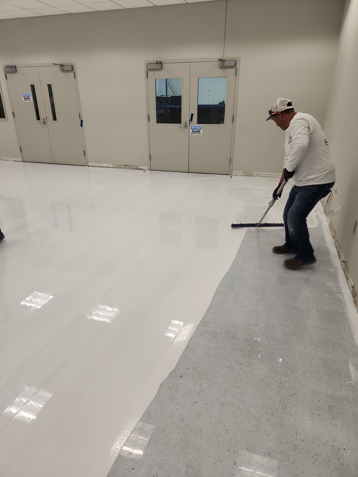 Worker applying white floor coating in a clean industrial room, with plastic-covered doors in the background.