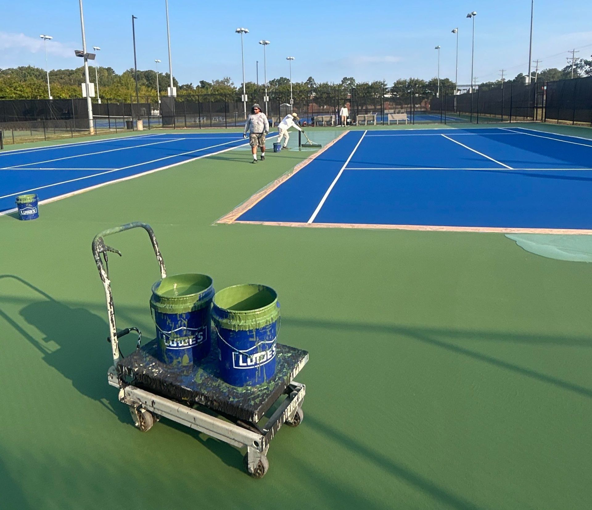 Paint cans on a cart in the foreground of a blue tennis court with players in the distance.