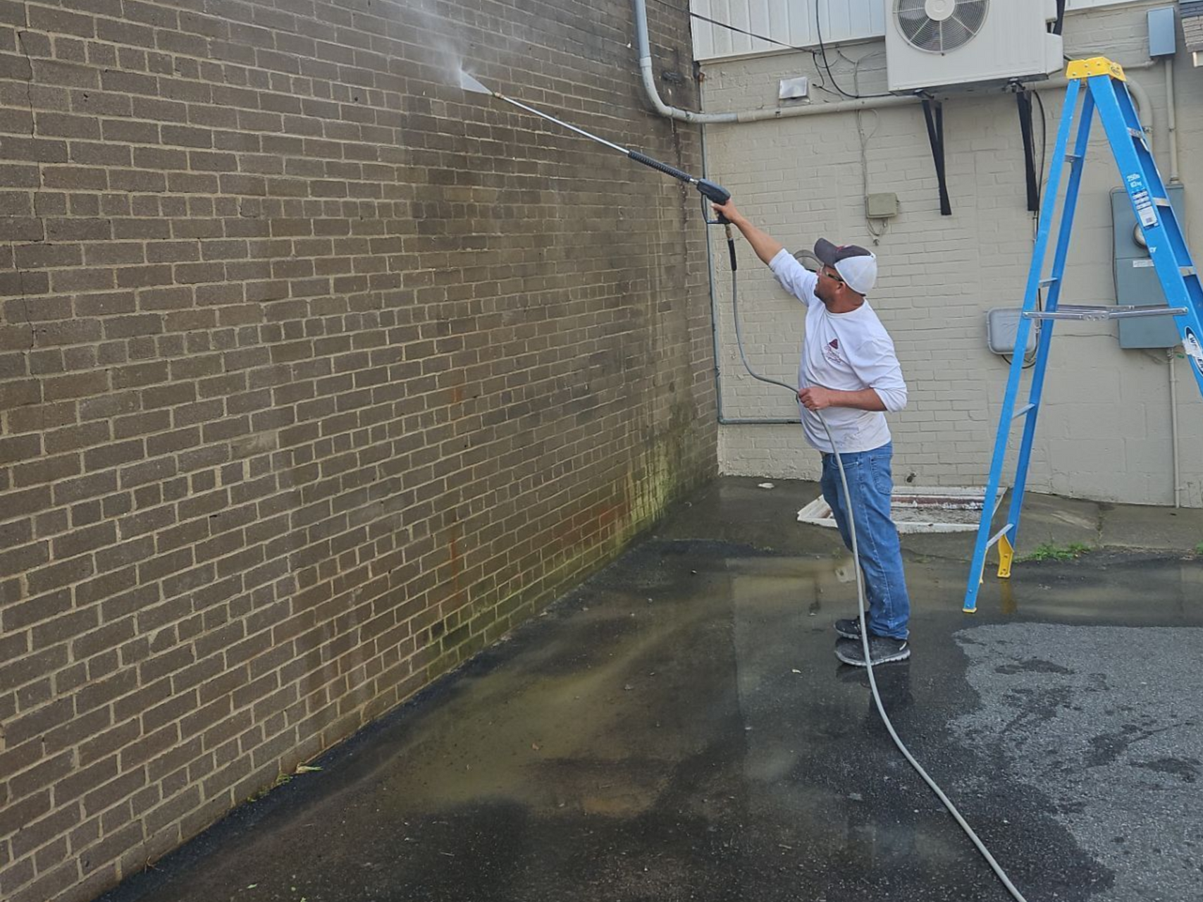Worker pressure-washing a brick wall beside a blue ladder in an outdoor alley