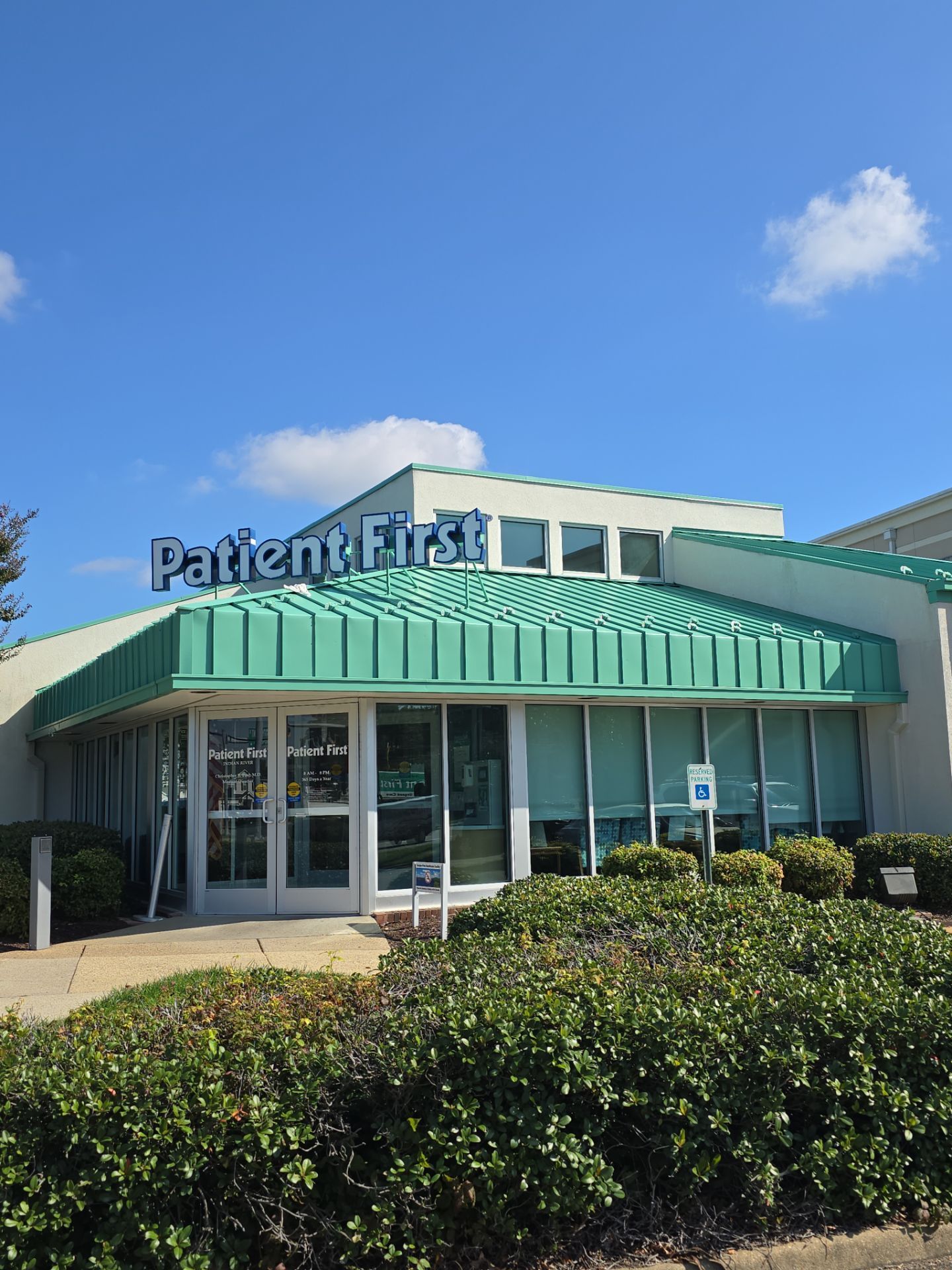 Patient First clinic exterior with green awning, glass entrance, and bushes under a blue sky