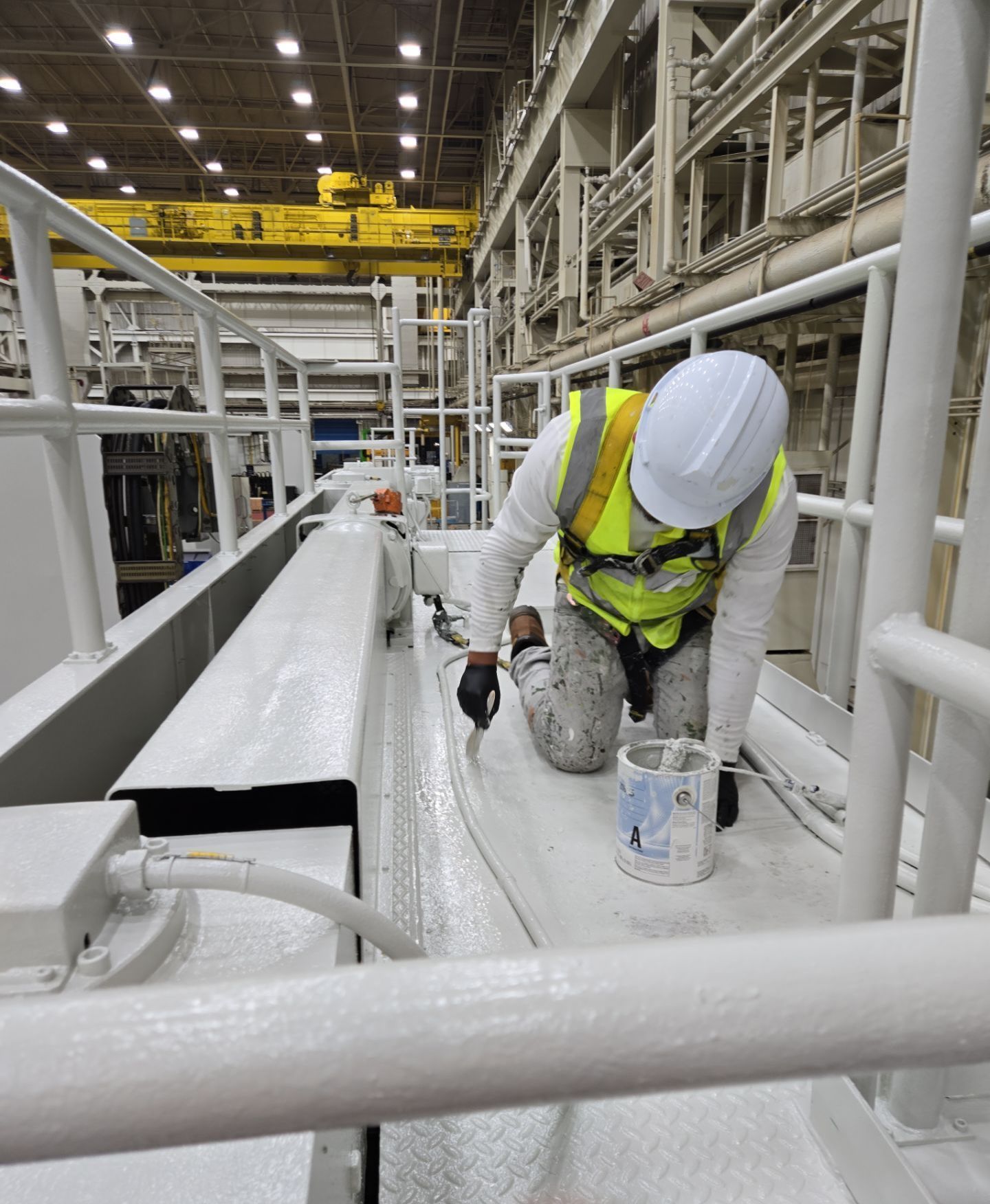 Worker in hard hat and safety vest painting a large industrial structure inside a factory facility