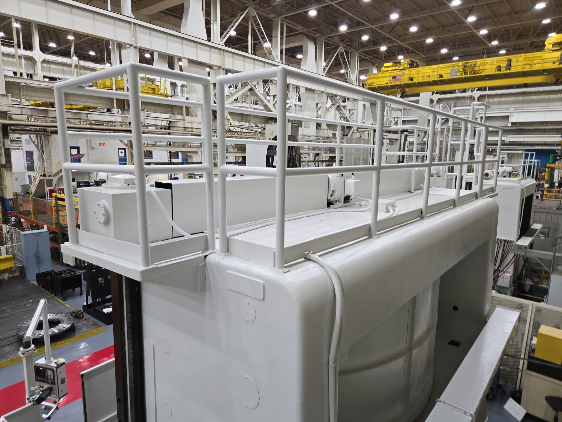 White industrial machine frame inside a factory, surrounded by overhead lights and workshop equipment.