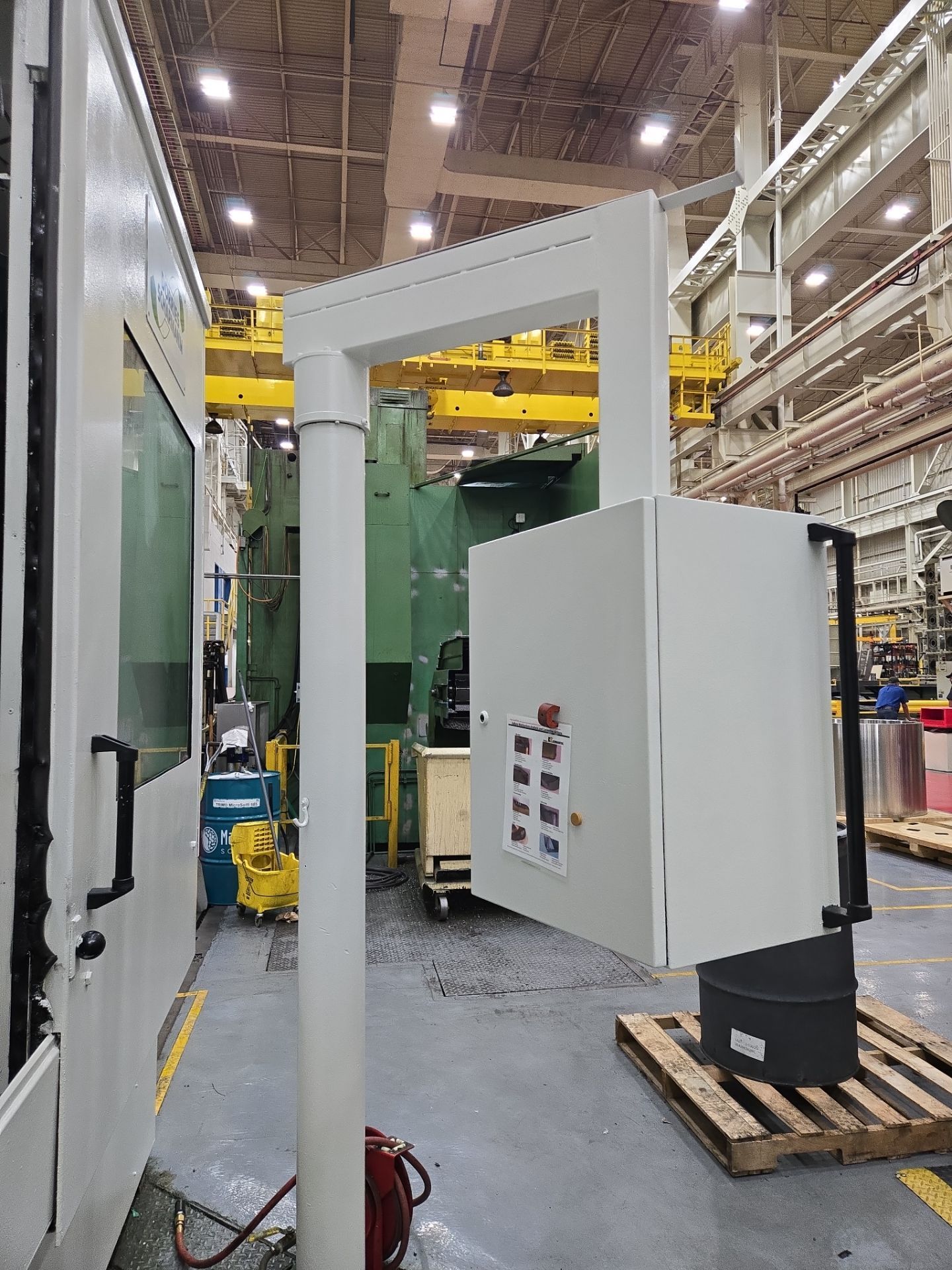 Large white industrial machine frame in a factory aisle, with control panel and overhead yellow crane.