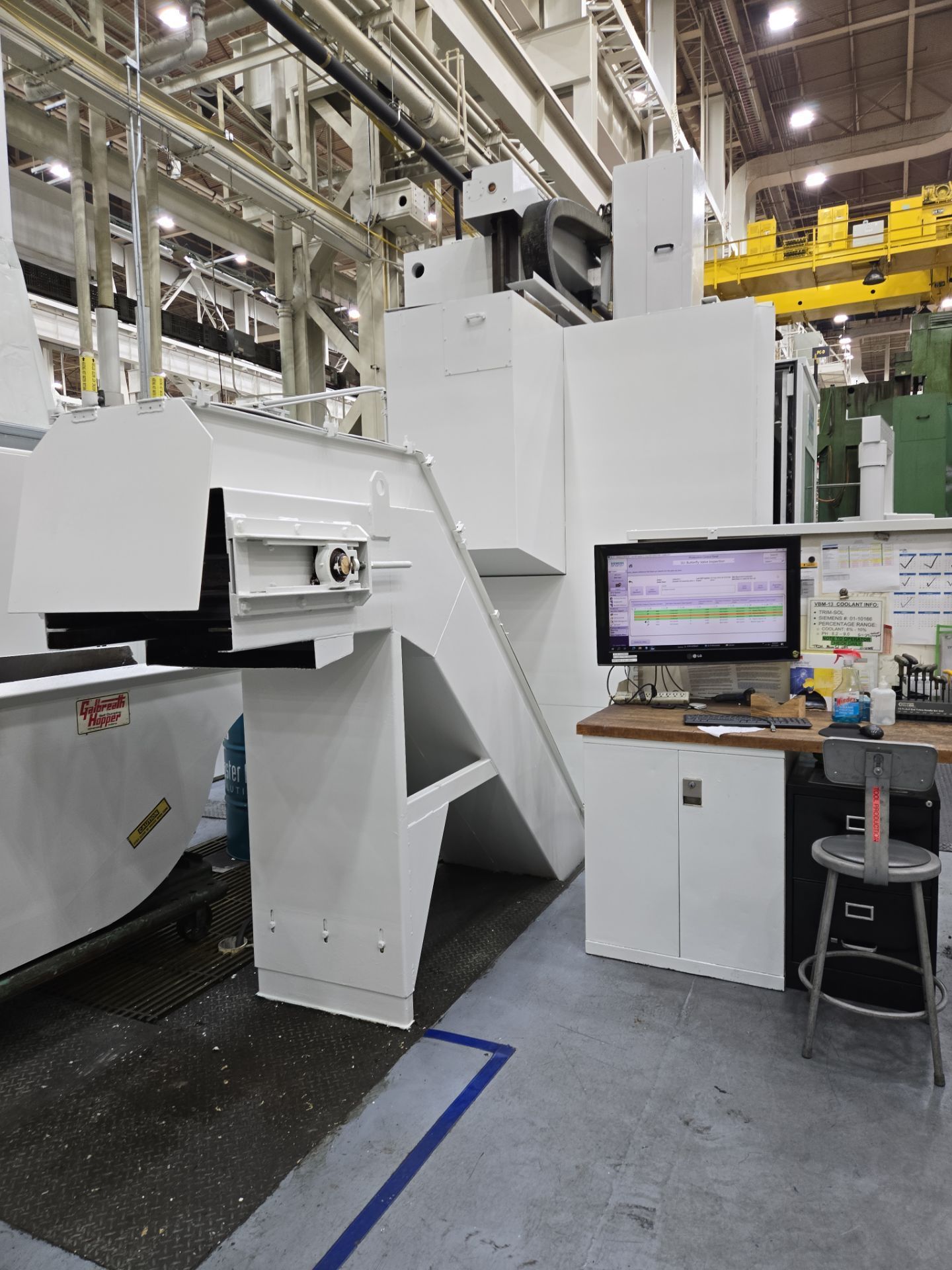 Industrial factory floor with a large white machine and a workstation monitor on a desk.