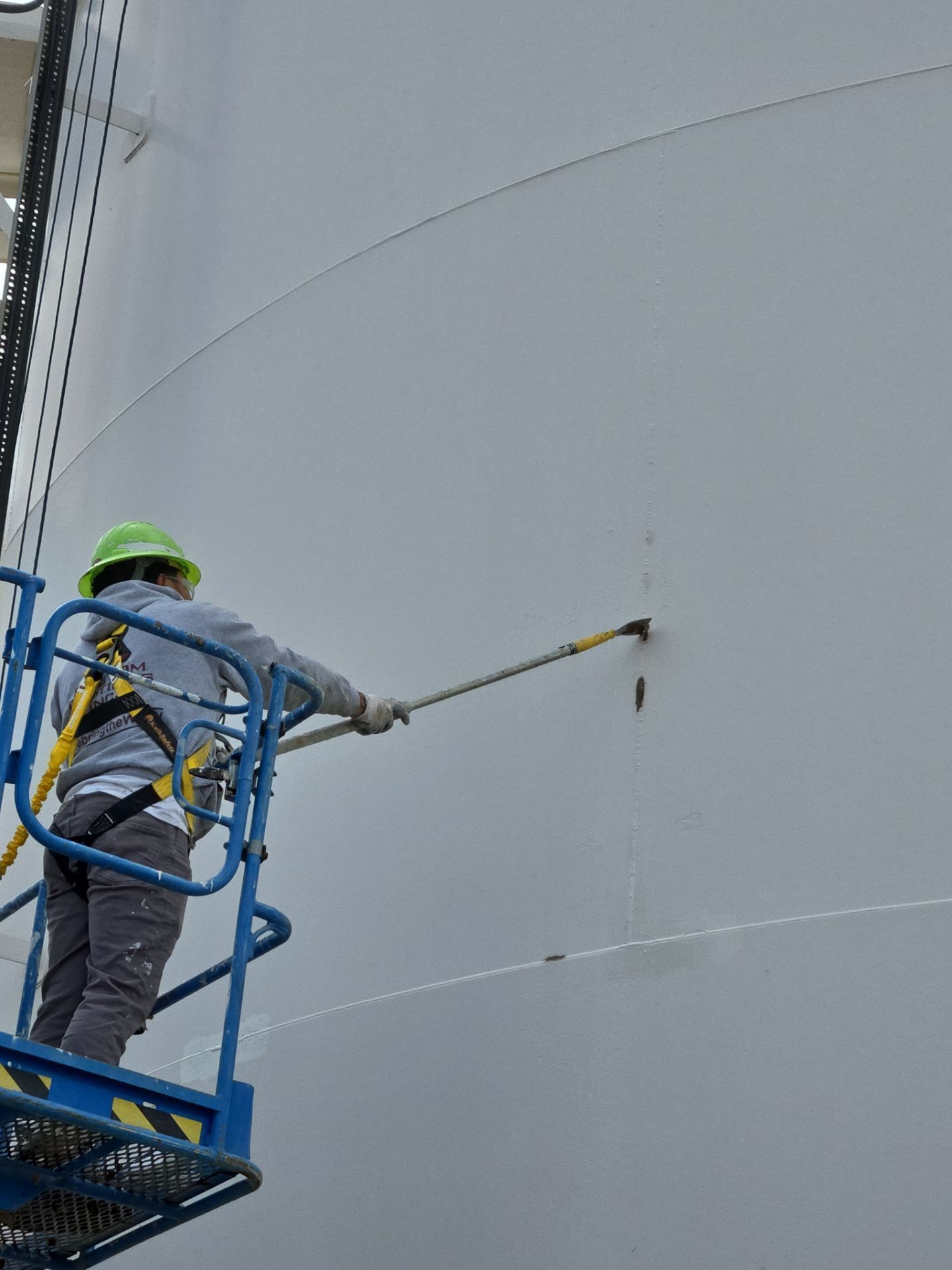 Worker in a blue lift pressure-washing a large gray tank wall