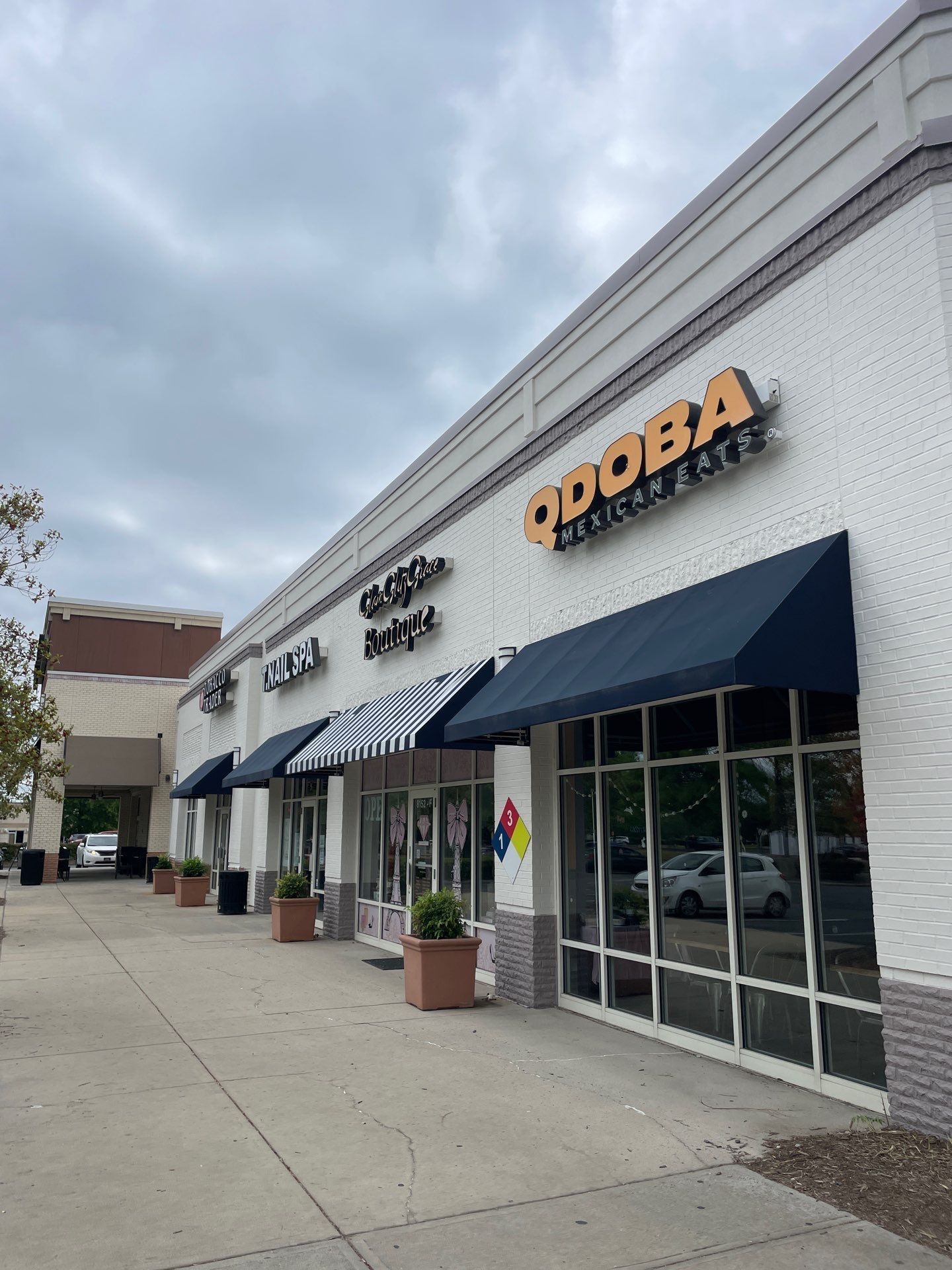 Strip mall storefronts with black awnings and a “QDOBA” sign under a cloudy sky