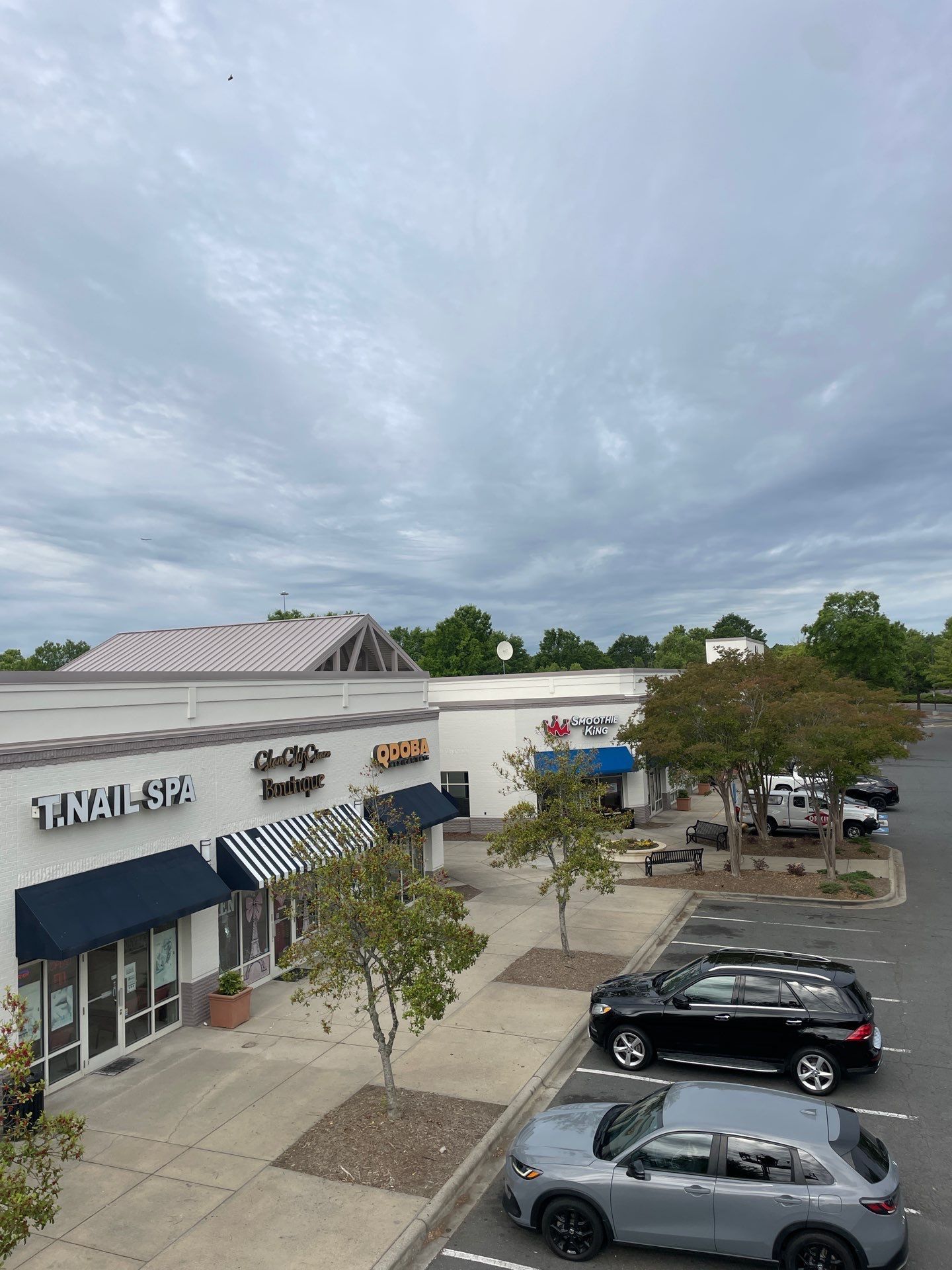 Strip mall with storefronts and parked cars under a cloudy sky