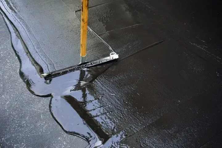 A squeegee with a yellow handle spreads thick, dark liquid sealant across a surface.