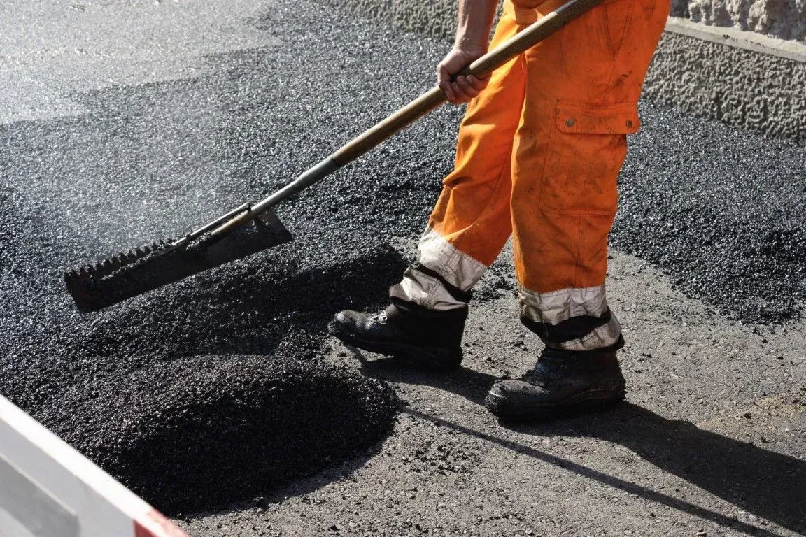 A worker in bright orange safety pants uses a rake to spread fresh, dark asphalt on a road surface.