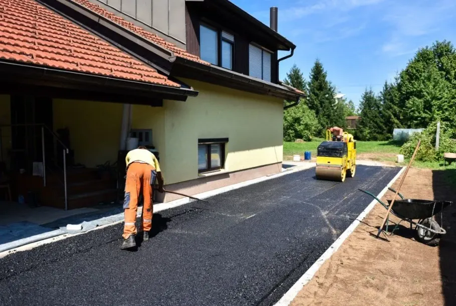 A construction worker works on a freshly paved asphalt driveway next to a house, with a steamroller in the background.