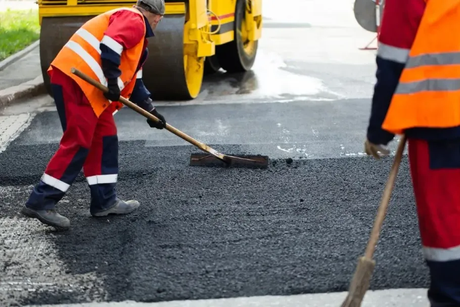 Two workers in high-visibility orange vests and red trousers spread hot asphalt on a road surface with a steamroller.