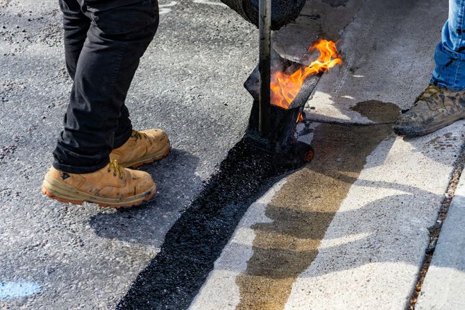 Two workers apply hot, liquid asphalt to seal a crack in a concrete pavement surface using a handheld torch.