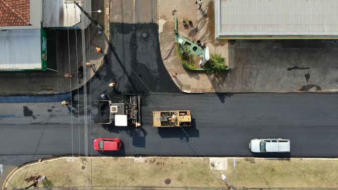 An aerial view of a road construction site featuring a paving machine, a yellow roller, and two cars on fresh asphalt.